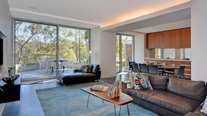 A modern living room with a large window, a sofa, a coffee table, and a dining area with a kitchen in the background. - Treehouse, Vincentia - 5 min walk to the beach (Vincentia)