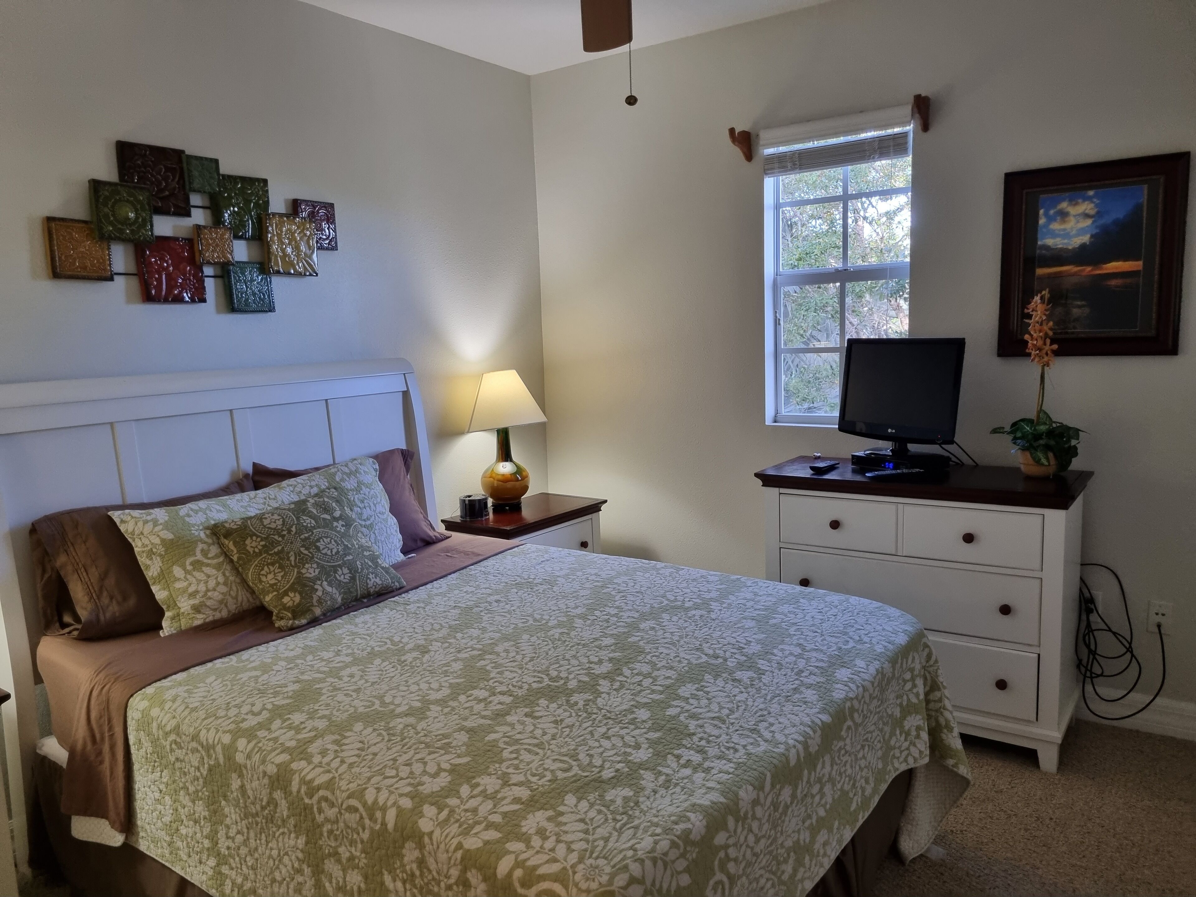 A bedroom with a bed, bedside lamp, TV, dresser, and a window with a view of trees.