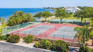 Camp de tennis amb vista al mar, envoltat de vegetació i un edifici blanc.