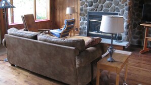 Un salon chaleureux avec une cheminée en pierre, des murs en bois, un canapé en cuir, un fauteuil à rayures bleues et une lampe posée sur une table en bois.