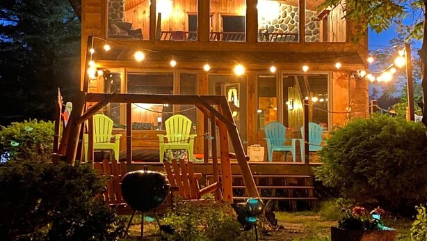 Une cabane en bois à deux étages, dotée d’une véranda, de nombreuses fenêtres et d’un espace extérieur aménagé pour s’asseoir.