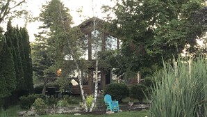 Une jetée en bois mène à un étang où se trouve un barbecue abrité, le tout étant entouré d’une végétation luxuriante et agrémenté d’une chaise bleue.