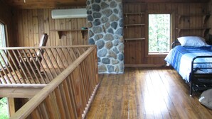 Une cabane en bois avec un mur en pierre, un lit recouvert d’une couverture bleue et une terrasse en bois.