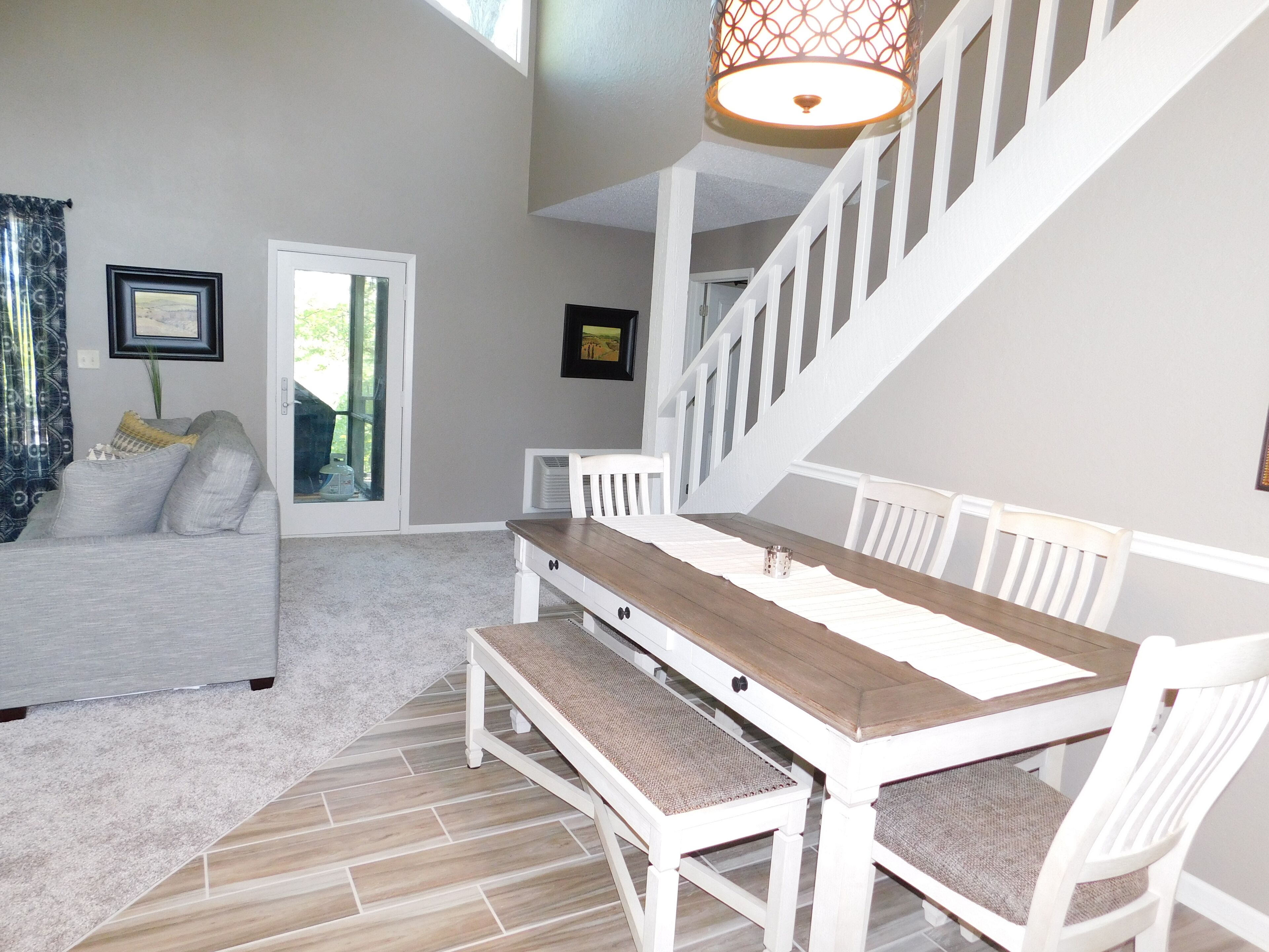Un salon spacieux avec un canapé blanc, une table à manger en bois avec des chaises blanches, un escalier et une grande fenêtre avec des rideaux.
