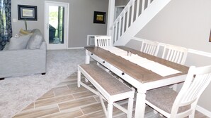 A spacious living room with a white sofa, a wooden dining table with white chairs, a staircase, and a large window with curtains.
