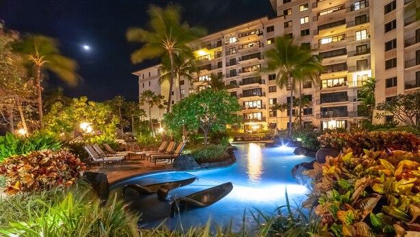 Une zone de piscine d’un complexe hôtelier, aménagée pour la nuit, avec des chaises longues et un grand bâtiment en arrière-plan.