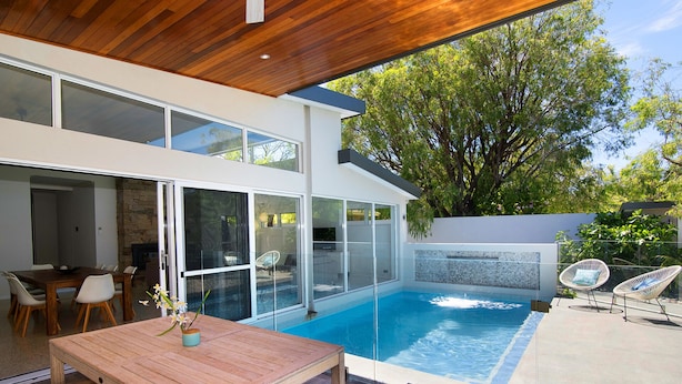 A modern outdoor pool area with a wooden table and chairs, a glass-enclosed patio, and a stone wall. - SALT - Luxury Oceanside Oasis (West Busselton)