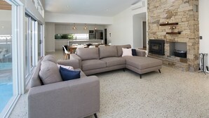 A modern living room with a stone fireplace, a large sectional sofa, and a kitchen in the background. - SALT - Luxury Oceanside Oasis (West Busselton)