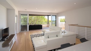 A modern living room with a white sofa set, a flat-screen TV, and a large sliding glass door leading to a balcony with a view of greenery. - SALT - Luxury Oceanside Oasis (West Busselton)