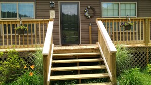 Una terraza de madera con una escalera que lleva a una puerta marrón, rodeada de follaje y plantas en macetas.