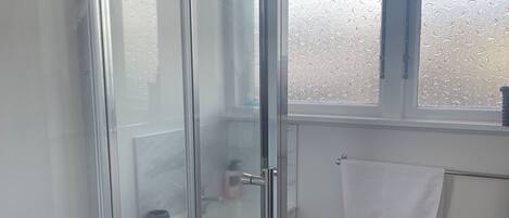 A bathroom with a glass shower enclosure, a white sink, and a window with a towel hanging on the wall.