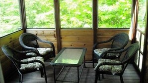 A covered porch with four wicker chairs and a small table, large windows offering a view of greenery. - Romantic getaway, private cabin overlooking Sawyer Lake  WIFI, TV, AC, Heat (Gilmanton)