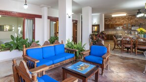 Un grand salon avec un canapé bleu, des chaises en bois, une table basse et une cuisine en arrière-plan.