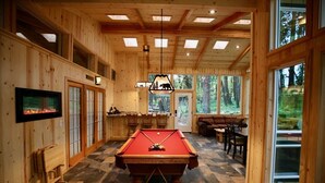 Ein Holzinnenraum mit Pooltisch, Bar und großen Fenstern mit Blick auf den Wald.