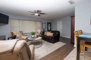 A living room with a brown leather sofa, a beige armchair, a coffee table, and a dining area with a wooden table and chairs.