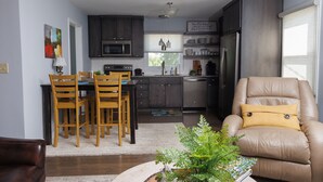 A modern kitchen with dark cabinets, a dining area with wooden chairs, and a beige armchair with a yellow pillow. - Cozy and Conveniently located close to KCI (KCMO)