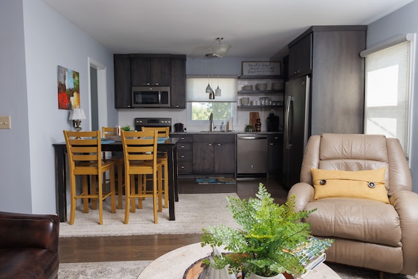 A modern kitchen with dark cabinets, a dining area with wooden chairs, and a beige armchair with a yellow pillow.