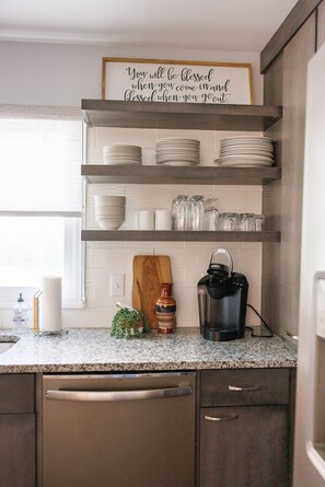 A kitchen with white cabinets, a granite countertop, a coffee maker, and a sign above the shelves.