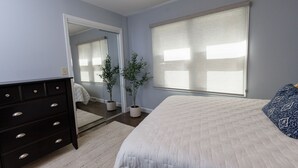 A bedroom with a bed, dresser, mirror, potted plants, and a window with blinds. - Cozy and Conveniently located close to KCI (KCMO)