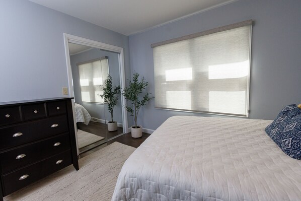 A bedroom with a bed, dresser, mirror, potted plants, and a window with blinds.