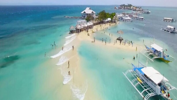 Vue aérienne d’une plage aux eaux turquoise et limpides, bordée d’un rivage sablonneux et ponctuée de plusieurs bateaux.
