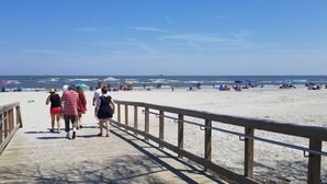 Une promenade en bois mène à une plage de sable où l’on trouve des parasols et des personnes qui se prélassent au soleil.