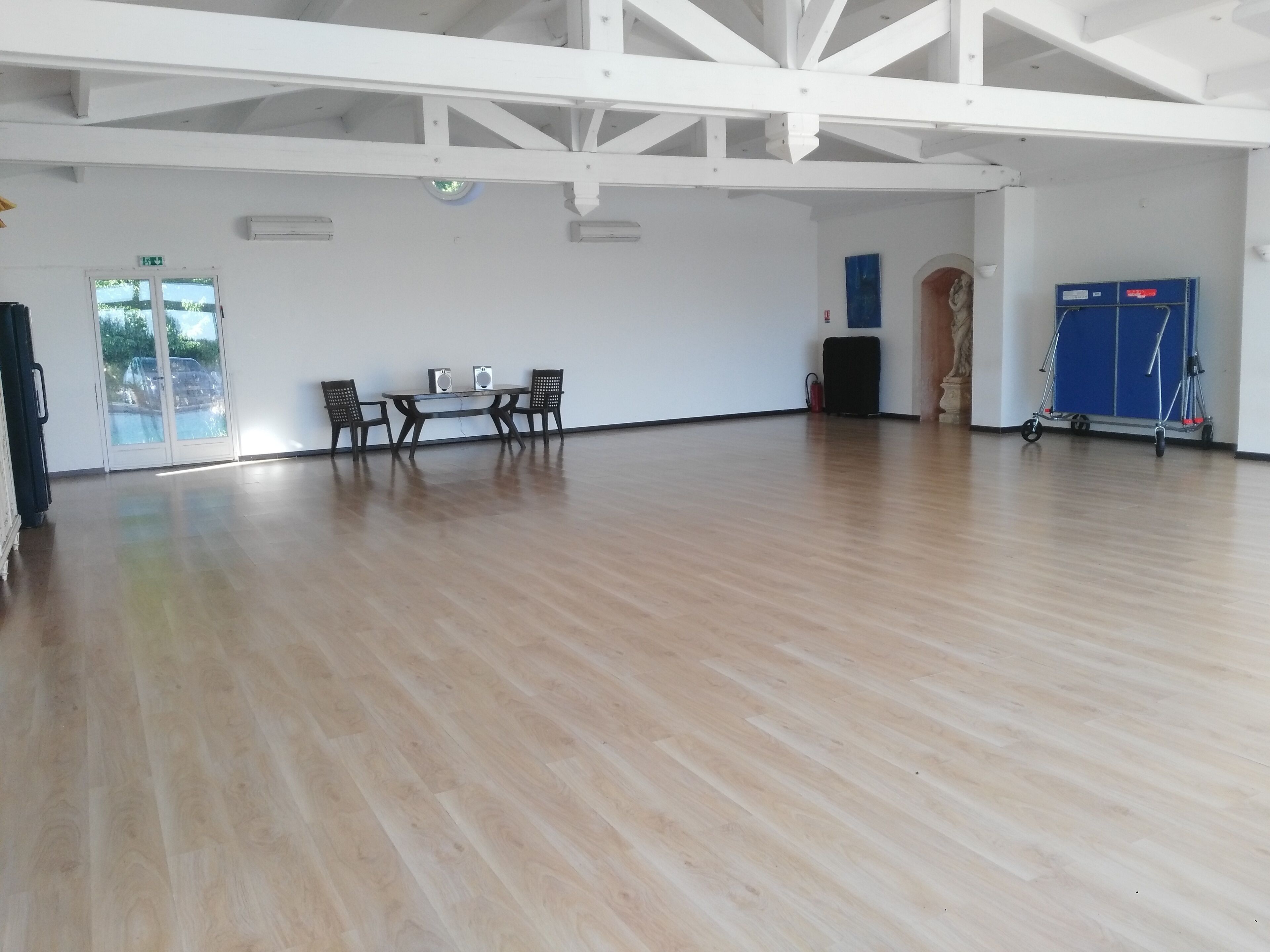 A spacious room with a wooden floor, white walls, and a high ceiling with exposed beams. There is a table with chairs, a blue cabinet, and a fire extinguisher on the wall.