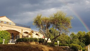 A historic building with arches and a balcony, surrounded by greenery and a rainbow.