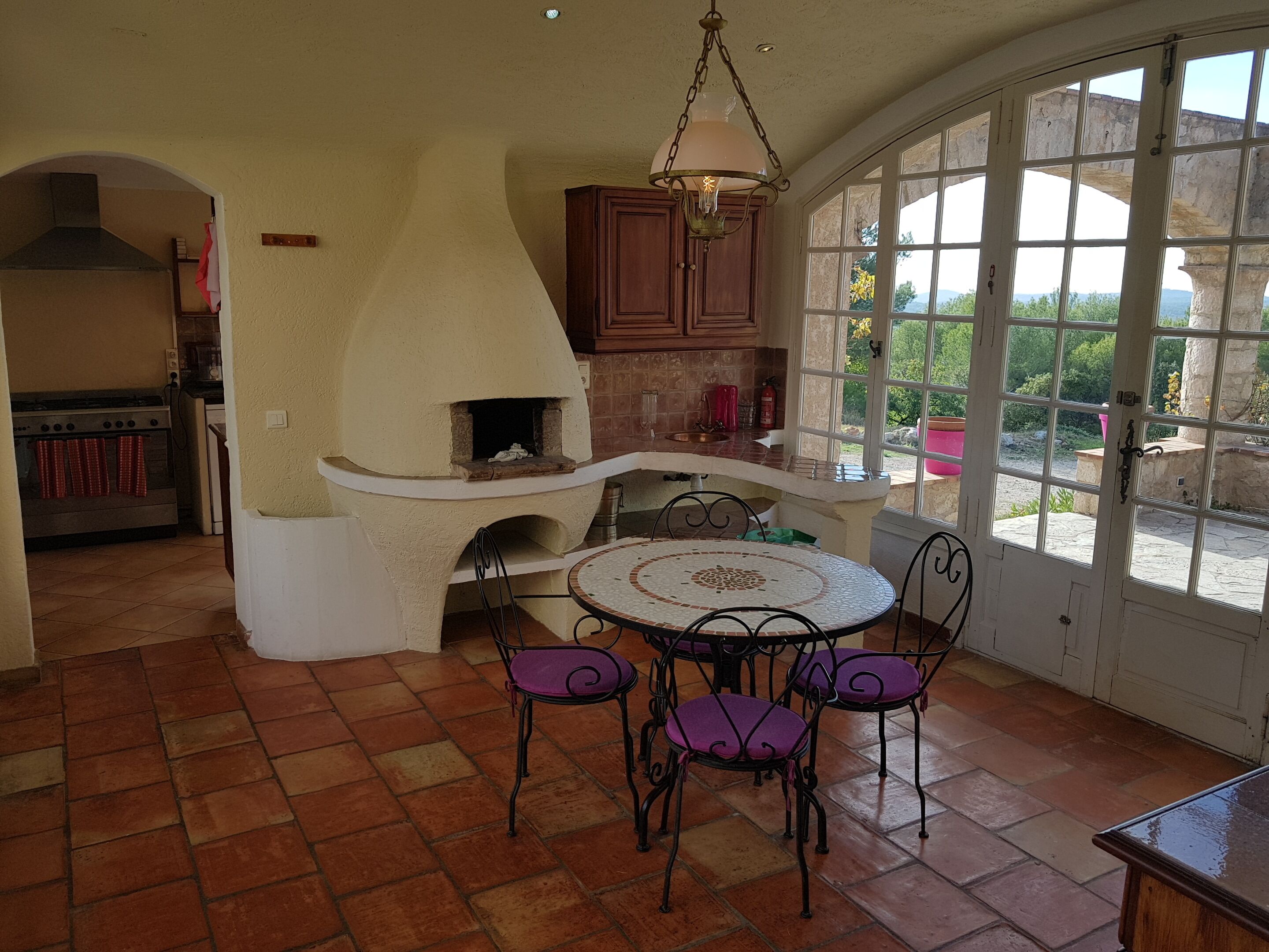Une cuisine dotée d’un four en pierre, une table à manger avec des chaises, et une vue dégagée grâce à de grandes fenêtres.