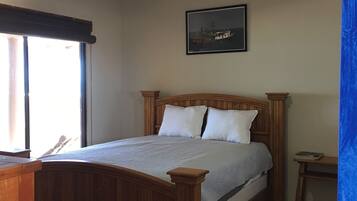 Une chambre avec un lit en bois, un ventilateur de plafond, un tableau encadré au mur et une fenêtre avec des stores.