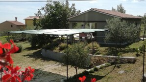Zona residencial con una casa, un espacio de estar techado al aire libre y un jardín con árboles y arbustos pequeños.