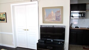 Una habitación con una puerta blanca cerrada, un mueble de entretenimiento negro, un cuadro de un edificio y una zona de cocina con microondas.