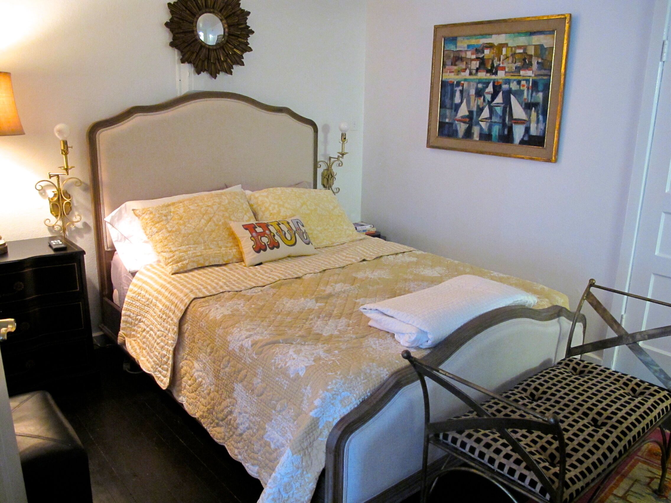 Una cama bien hecha con un edredón y almohadas, una mesita de noche con una lámpara, una silla con asiento de mimbre y un cuadro enmarcado en la pared.