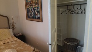 Un dormitorio con cama, una mesita de noche con libros, un perchero, una cesta de mimbre y un ventilador blanco.