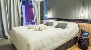 A neatly made bed with a white comforter and two pillows, a bedside table with a phone and a small screen, and a window with curtains. - Plaisance.  A 160 m2 apartment with exceptional view. (Athens)