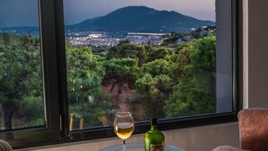 Een kamer met een groot raam dat uitkijkt over een stadsgezicht en bergen. Er staat een glazen tafel met een fles en een glas wijn. Twee gestreepten stoelen zijn zichtbaar.