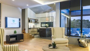 Een moderne woonkamer met een televisie, gestreepte banken, een ronde salontafel en uitzicht op de keuken en het balkon.