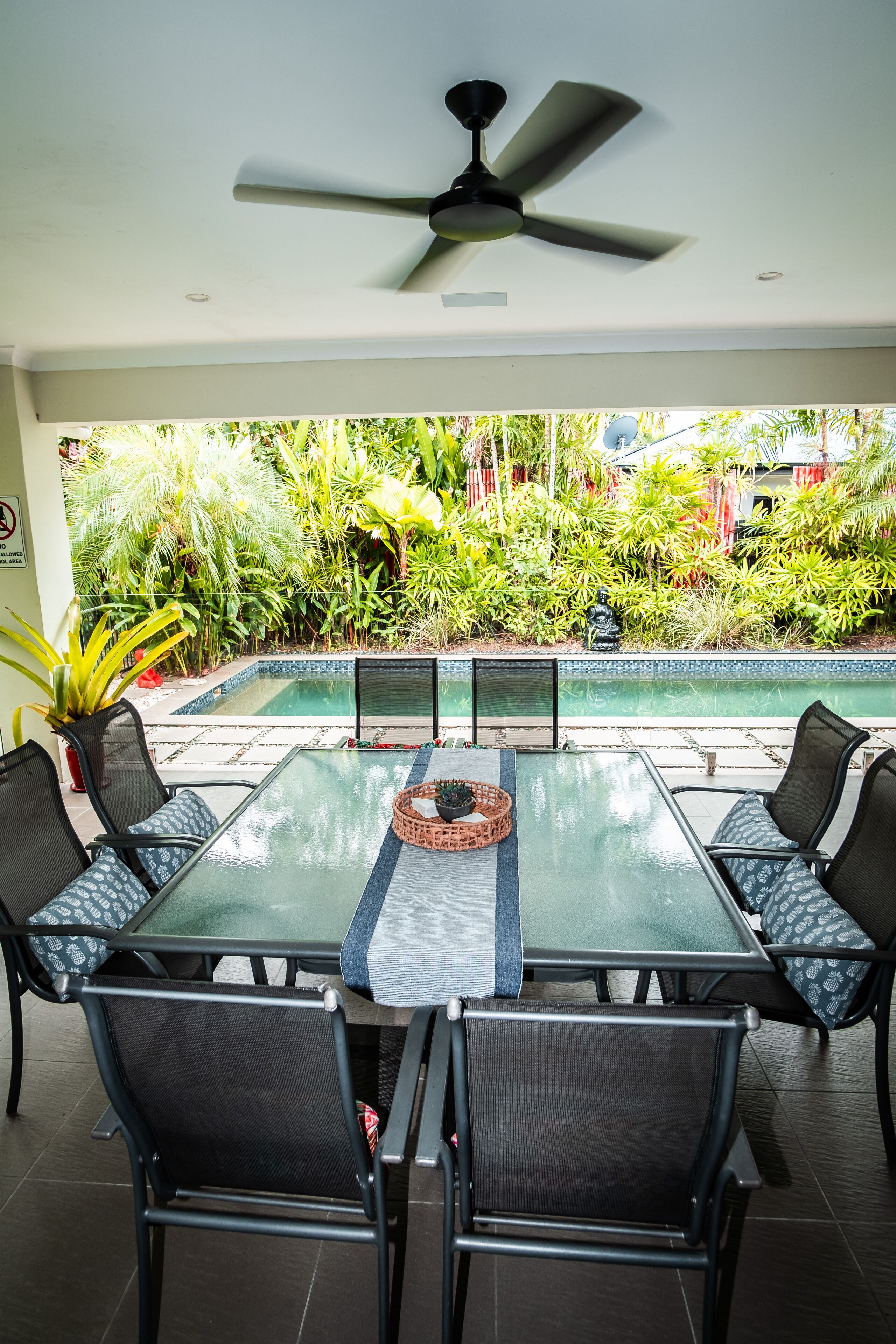 Una mesa de comedor con cubierta de vidrio, cuatro sillas, un ventilador de techo y vistas a una piscina y abundante vegetación.