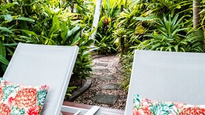 Eine Terrasse mit zwei weißen Liegestühlen, jeweils mit Blumenkissen. Die Terrasse ist von üppigen tropischen Pflanzen und einem Steinweg umgeben.