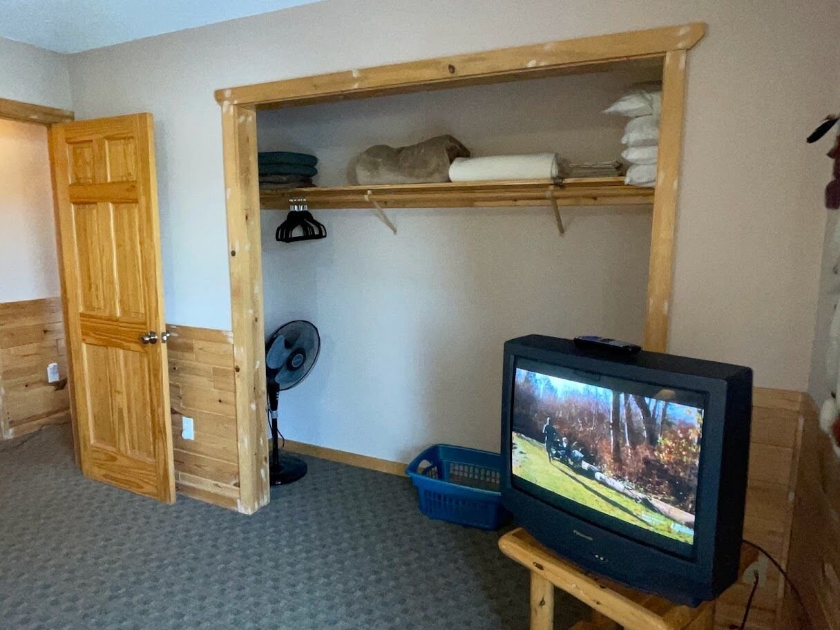A room with a wooden wardrobe, a television on a wooden stand, and a blue plastic basket.