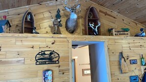 A wooden cabin interior with a deer head mounted on the wall, a wooden sign with a mountain and river, and a door leading to another room.