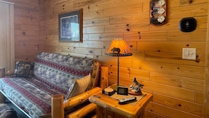 A cozy cabin interior with a wooden log wall, a sofa with patterned cushions, a table with a lamp and remote controls, and a clock.