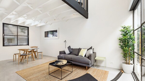 A modern living room with a grey sofa, wooden chairs, a coffee table, and a large window with blinds.