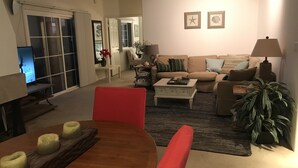 Ein Wohnzimmer mit einer Couch, einem Couchtisch und einem Fernseher. Es gibt einen Esstisch mit Stühlen und einer Pflanze.