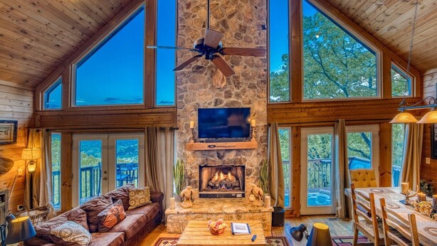 A spacious interior with a stone fireplace, wooden ceiling, and large windows offering views of the outdoors.