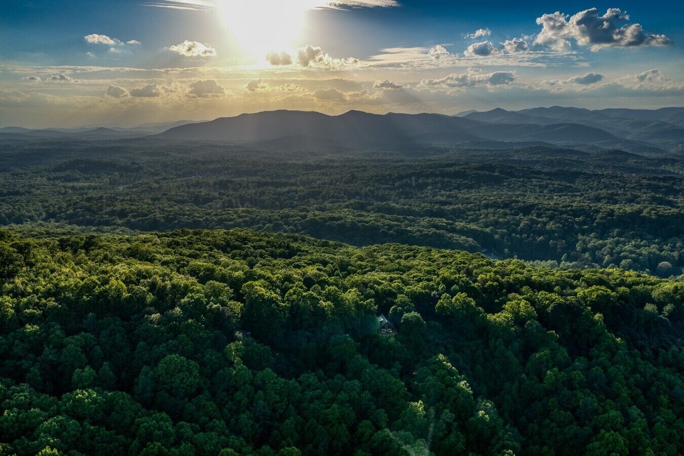 Un paysage boisé, avec une chaîne de montagnes en arrière-plan et une maison au premier plan.