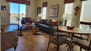 A living room with a leather sofa, a coffee table, and a dining table with chairs. There are windows with blinds, a door leading outside, and a view of the outside. - Up-scale 3 Room Suite Located In A Popular SW Resort At @2900 Feet Elevation (Green Valley)