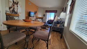 A dining area with a round wooden table, wicker chairs, and a bowl on the table. There is a television on a stand, a sofa, and a window with blinds. - Up-scale 3 Room Suite Located In A Popular SW Resort At @2900 Feet Elevation (Green Valley)