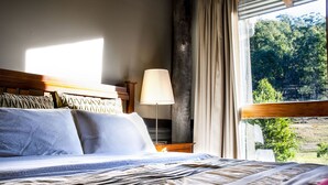 Una cama con una colcha a rayas, una lámpara de noche y vistas a un bosque a través de una ventana.