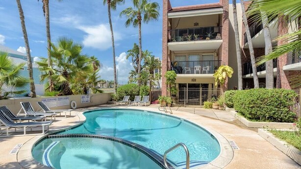 A pool area with lounge chairs, palm trees, and a two-story building with balconies. - Unit 101 Villa Del Sol (South Padre Island)